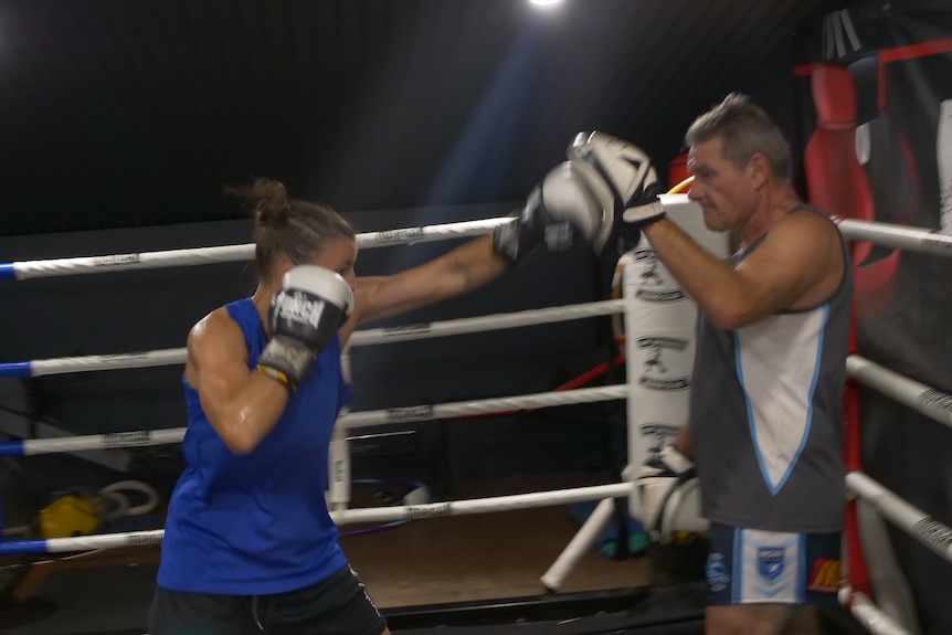 Jess in the ring with her coach punching up to his hand pad, one glove protecting her face.