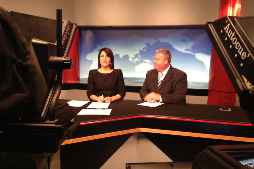 Jo Palmer and Rob Fairs sit behind a desk while reading the news. Jo is looking at the camera, Rob is looking at Jo.