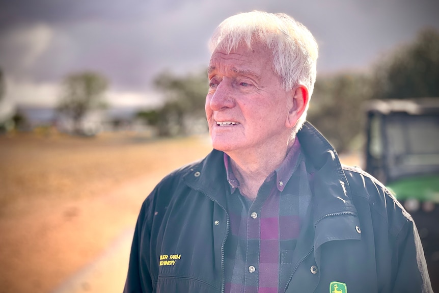 A man wearing a flannelette shirt and jacket stands in a paddock with a tractor in the background.