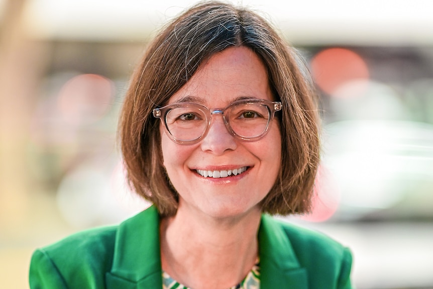 A headshot of a women with brown bobbed hair, glasses and green jacket.