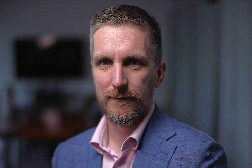 A man with a reddish-grey beard wearing a light pink business shirt and checkered blue suit jacket sits in a professional office
