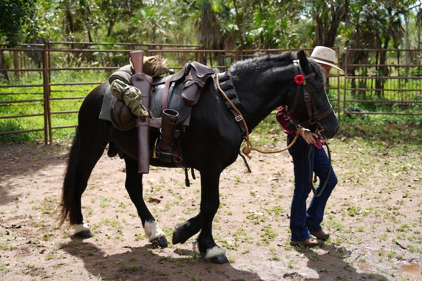 A woman leading a horse, with a red poppy tucked behind its ear, in a horse pen.