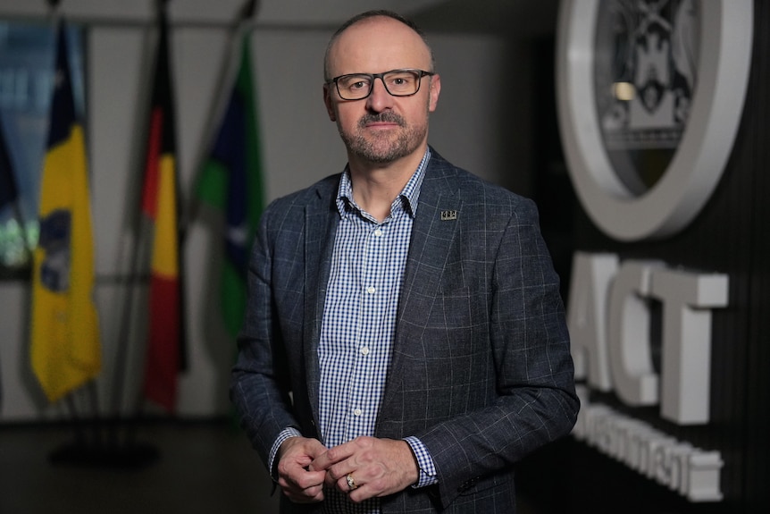 A man stands a looks at the camera, near flags and the ACT government logo.