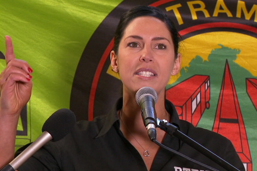 A woman with dark hair speaking into a microphone with her hand in the air.