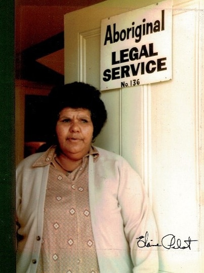 A woman with black hair stands in front of a doorway. 