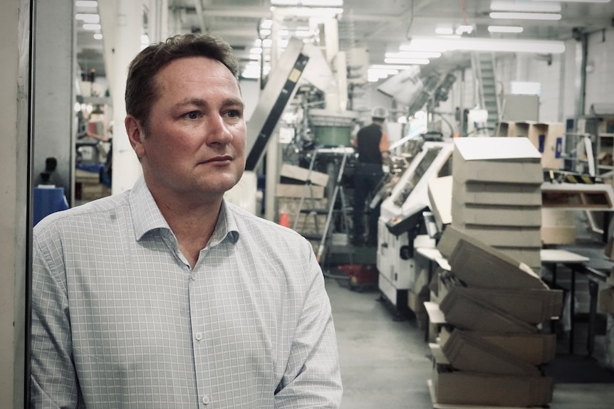 Aleks looks serious, stands in a factory with equipment, a man in plastic cap works behind.