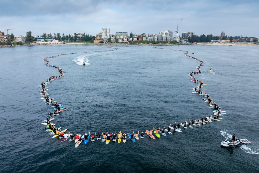 Image of a thousand surfers on their boards in the ocean. They're in a circle holding hands.