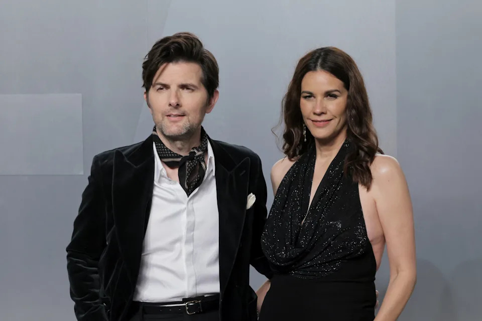 Adam Scott in a velvet suit with a scarf and Naomi Scott in a glittering halter dress pose on a formal event red carpet