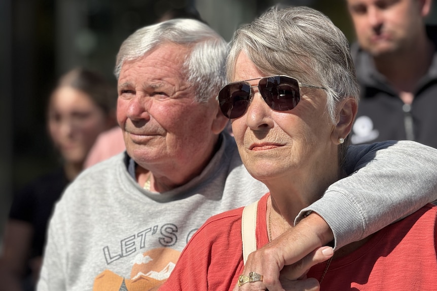 Two people with grey hair stand with their arms over each other.