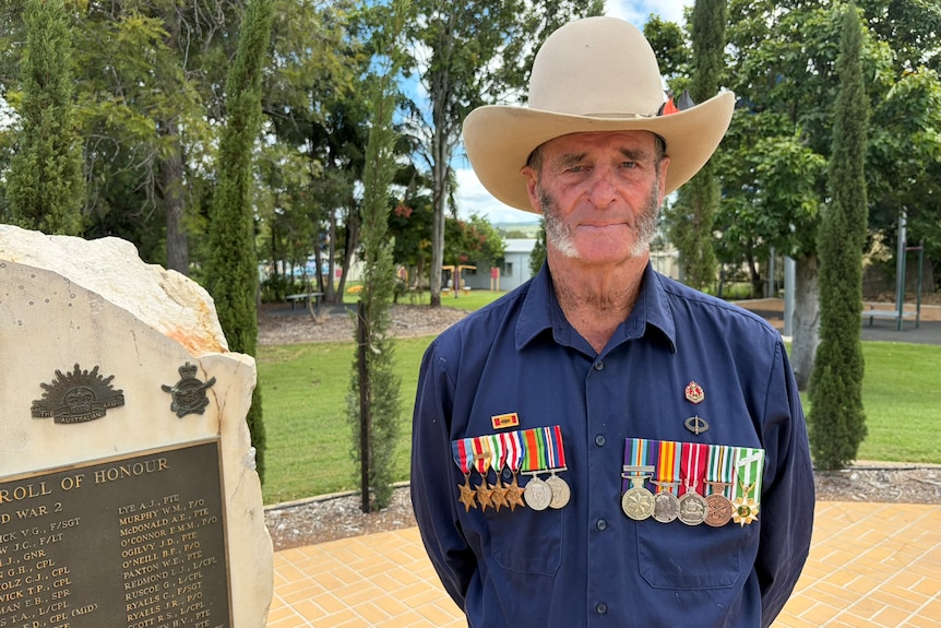 A man in a big hat with a row of medals on both sides of his chest standing next to a war monument