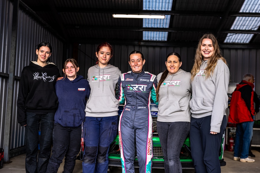 Imogen stands in the centre with five other women next to her and her car behind her.