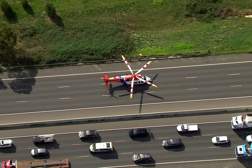 A red and white helicopter with a blue strip sits on a freeway as cars pass on the other side of the roadway.