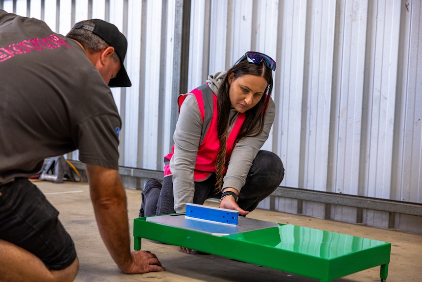 Natasha wears a high vis pink vest and kneels, working on a green car part.