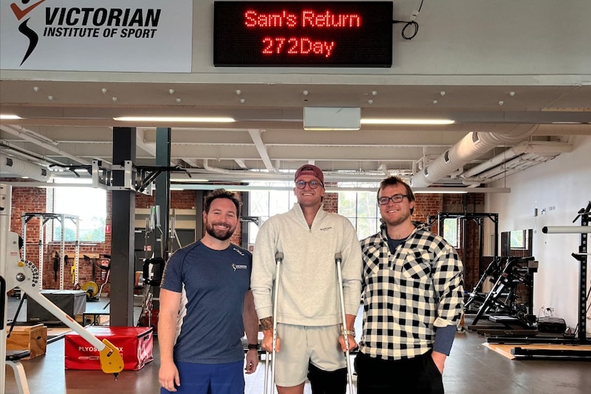 Three men stand in a gym, below a sign saying "Sam's return, 272 days'.