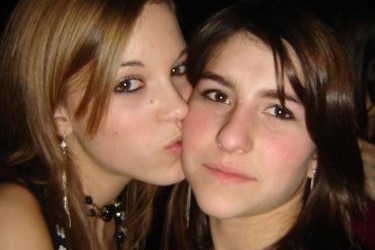 Two teenagers, one with dark blonde hair, the other with brown hair, stand cheek-to-cheek