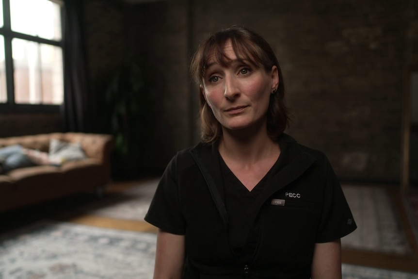 A woman with a short brown bob and fringe wearing black surgical scrubs sits in an industrial-style living room.