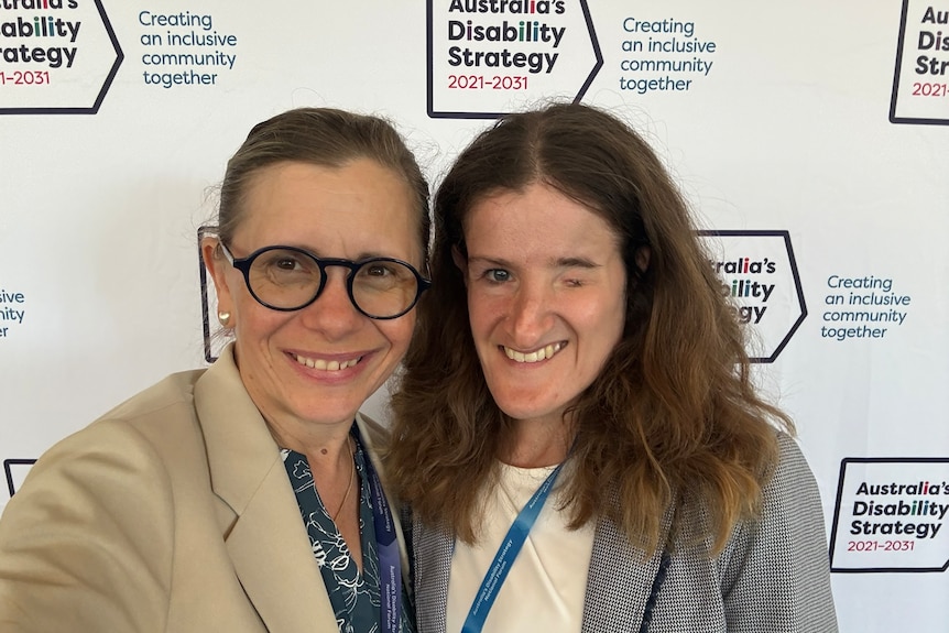 Two smiling, dark-haired women, one with facial difference, in front of a disability strategy backdrop.