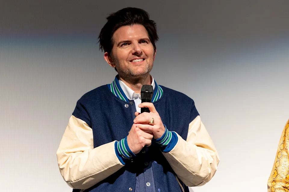Adam Scott smiling, wearing a varsity jacket, holding a microphone at a public event