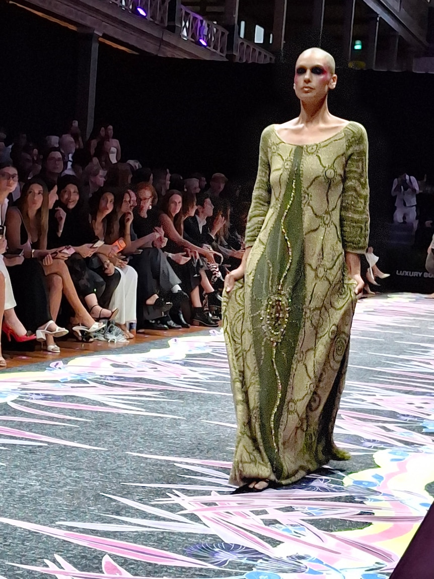 a woman is walking down a runway wearing a long knitted green dress with patterns. the model is balled and has red cheeks