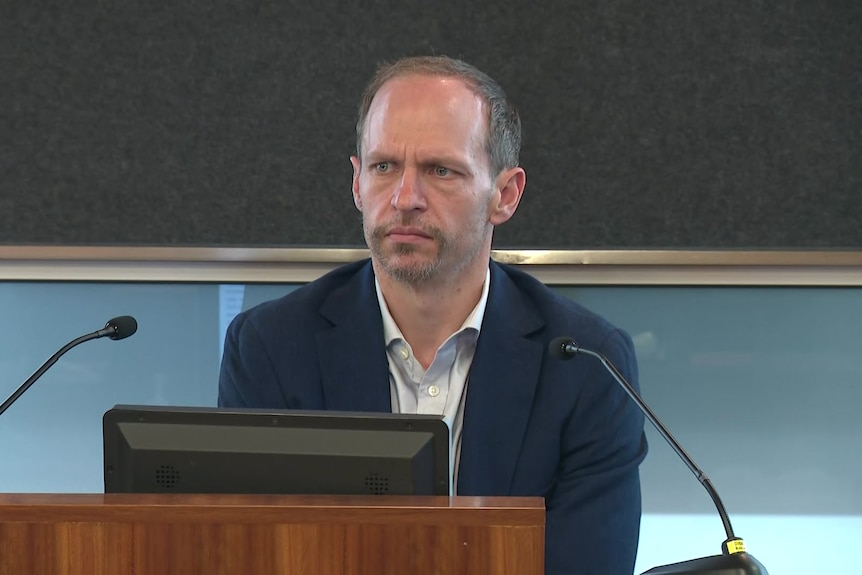 A man in a suit sitting in the dock at a parliamentary inquiry.