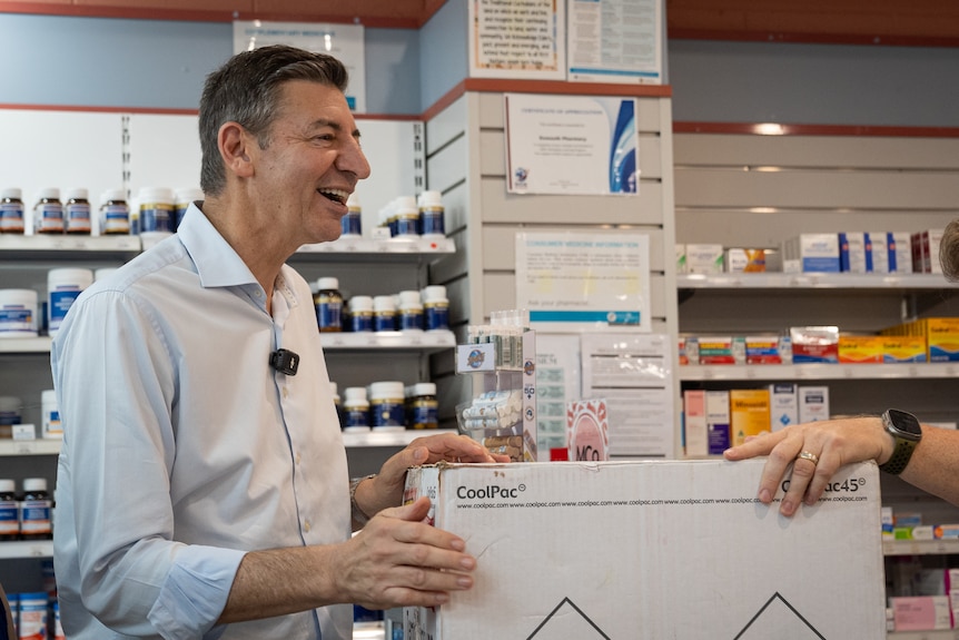 A middle-aged man with dark hair wears a business shirt and smiles while standing in a pharmacy.