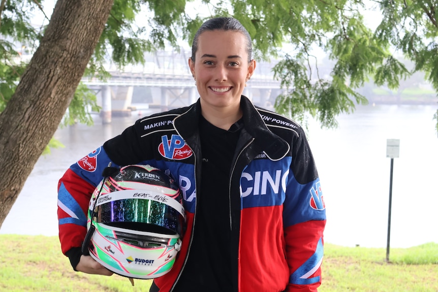 Imogen wears a red and blue jacket and holds her helmet, smiling with the river behind her.