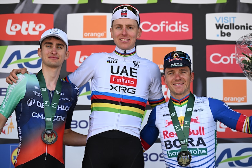 Paul Seixas (left), Tadej Pogačar (centre) and Remco Evenepoel (right) stand arm in arm wearing medals.