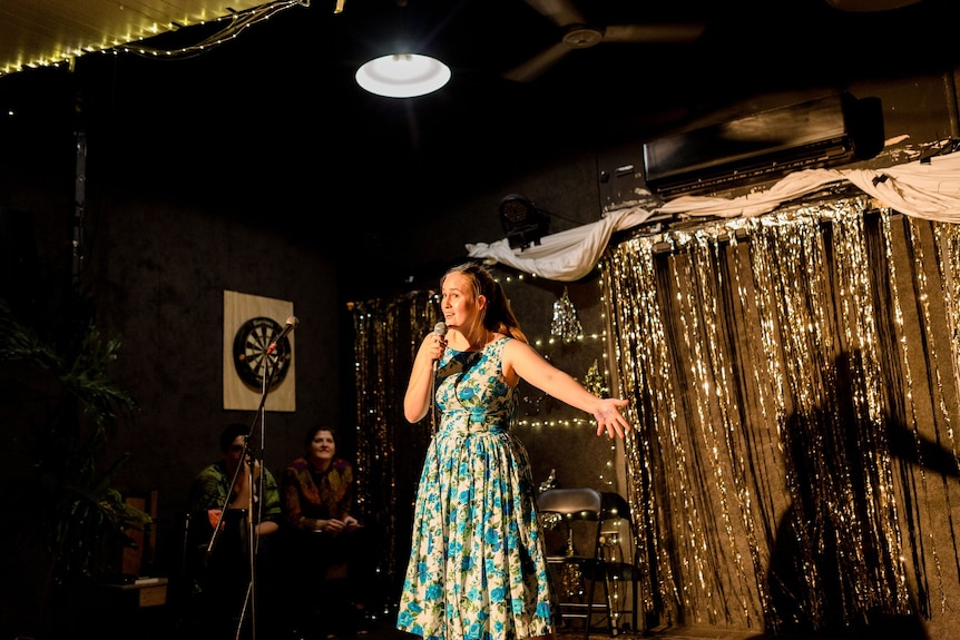 Grace Jarvis holds out an arm while performing standup in a retro dress against a tinsel backdrop.