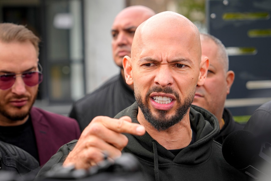 Andrew Tate angrily speaking outside court surrounded by people.