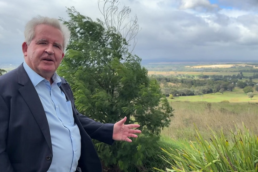 A man with white hair, wearing a blue shirt and blazer stands in front of a lookout 