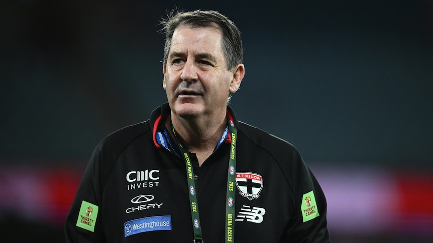 A St Kilda AFL coach stands on the ground looking downfield ahead of a match.
