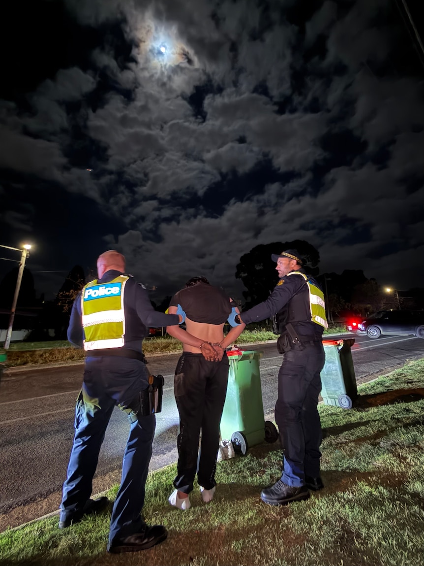 Two police officers in navy uniforms with bright yellow vests hold the arms of a man with a bare torso and dark pants at night.