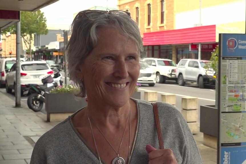 A older woman with short hair wearing a grey shirt and holding the brown strap of a handbag stands on a busy footpath.