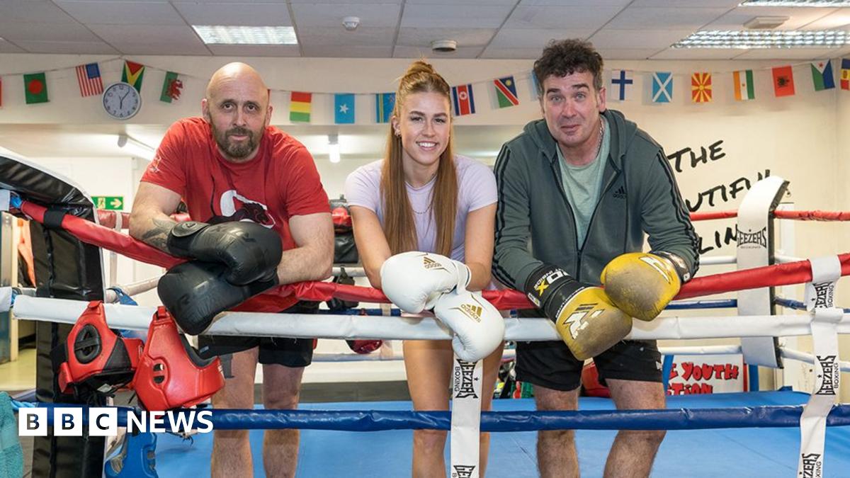 Three coaches stand facing the camera leaning on the ropes of a boxing ring wearing boxing gloves