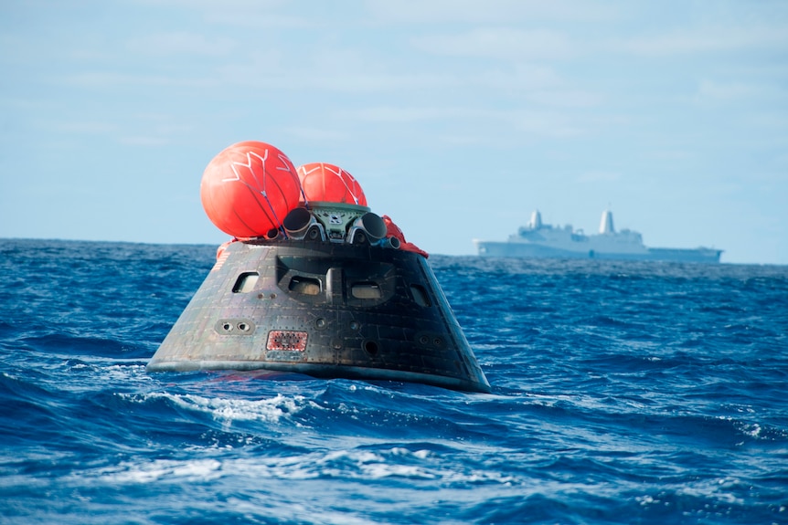 NASA Orion space capsule floats in the ocean with orange balloons and a navy ship in the background.