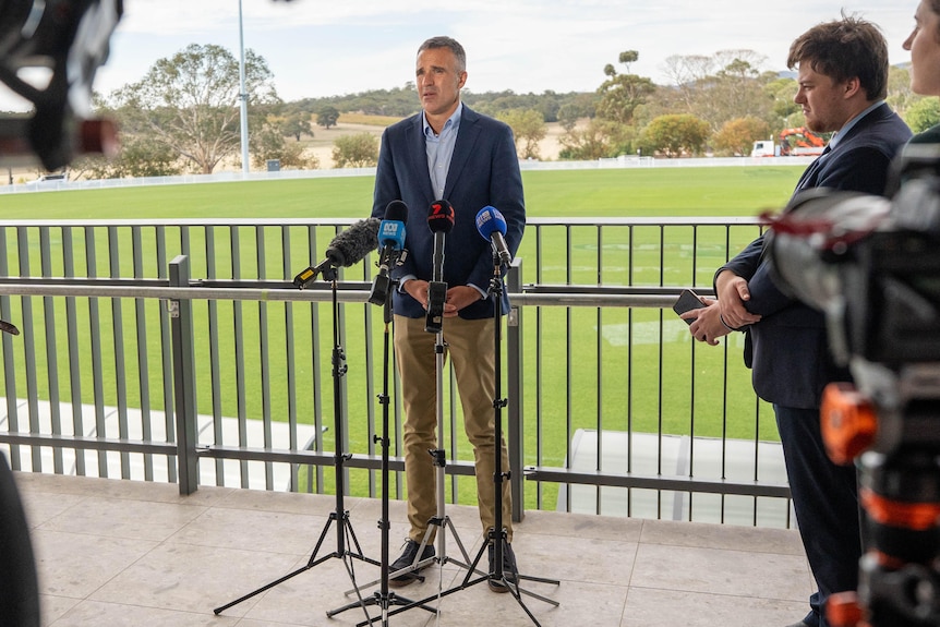 A man wearing a suit jacket and collared shirt in front of a football oval