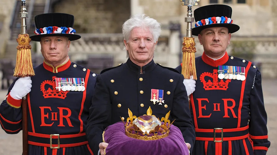 Former pageant master Bruno Peek standing in the middle of two Beefeaters. On a purple cushion, he is carrying a Diamond Jubilee 'Diamond'.
