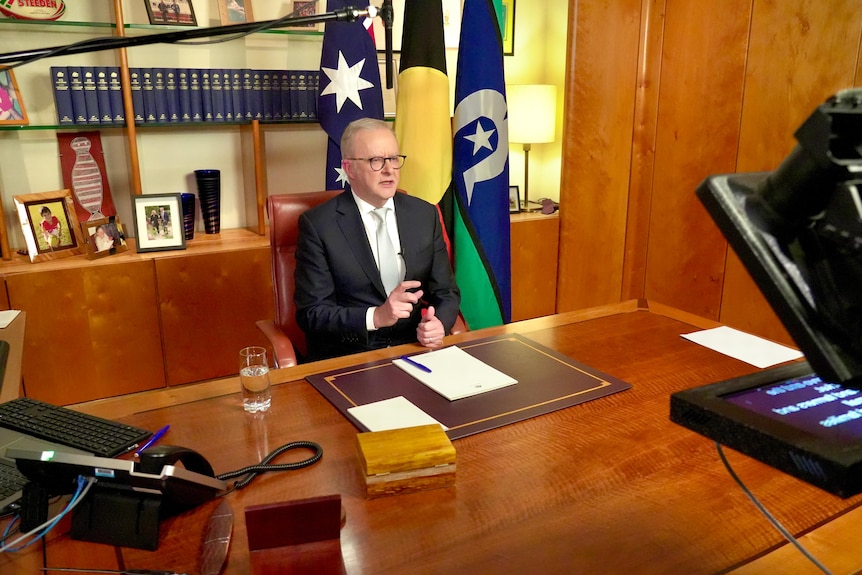 A man in a suit sits at a desk and reads a teleprompter, looking in to a camera.