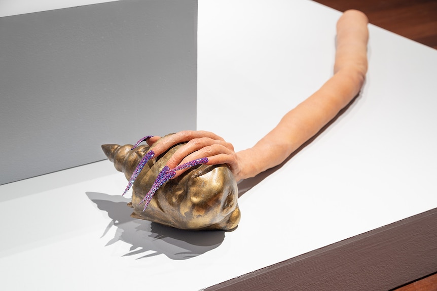 In a white gallery, on a platform rests an elongated arm, with a carefully purple-claw-manicured hand, holding a shell.