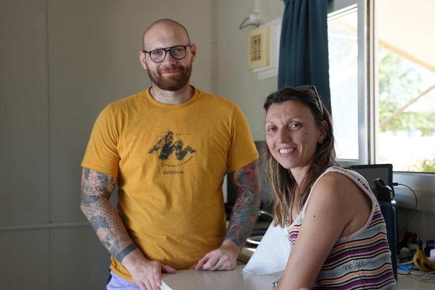 A smiling, bearded man with tattoos and a smiling, long-haired woman in a house.