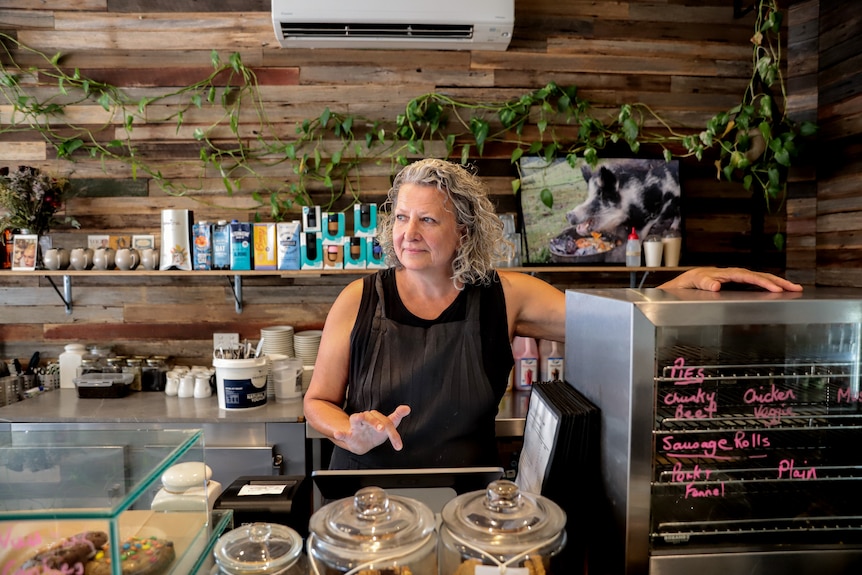 Woman with grey hair and black top stands behind cafe counter with hand over register looking out towards window 