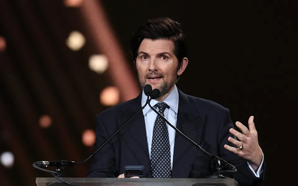 Adam Scott in a suit and polka dot tie speaks at a podium, gesturing with one hand