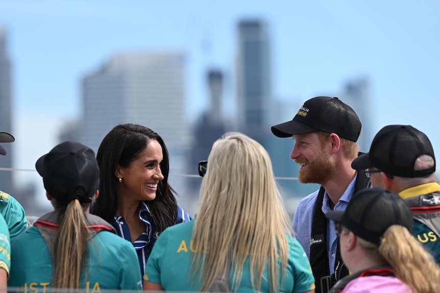 Harry and Meghan look at each other smiling in front of a group of people with the cityscape behind them.