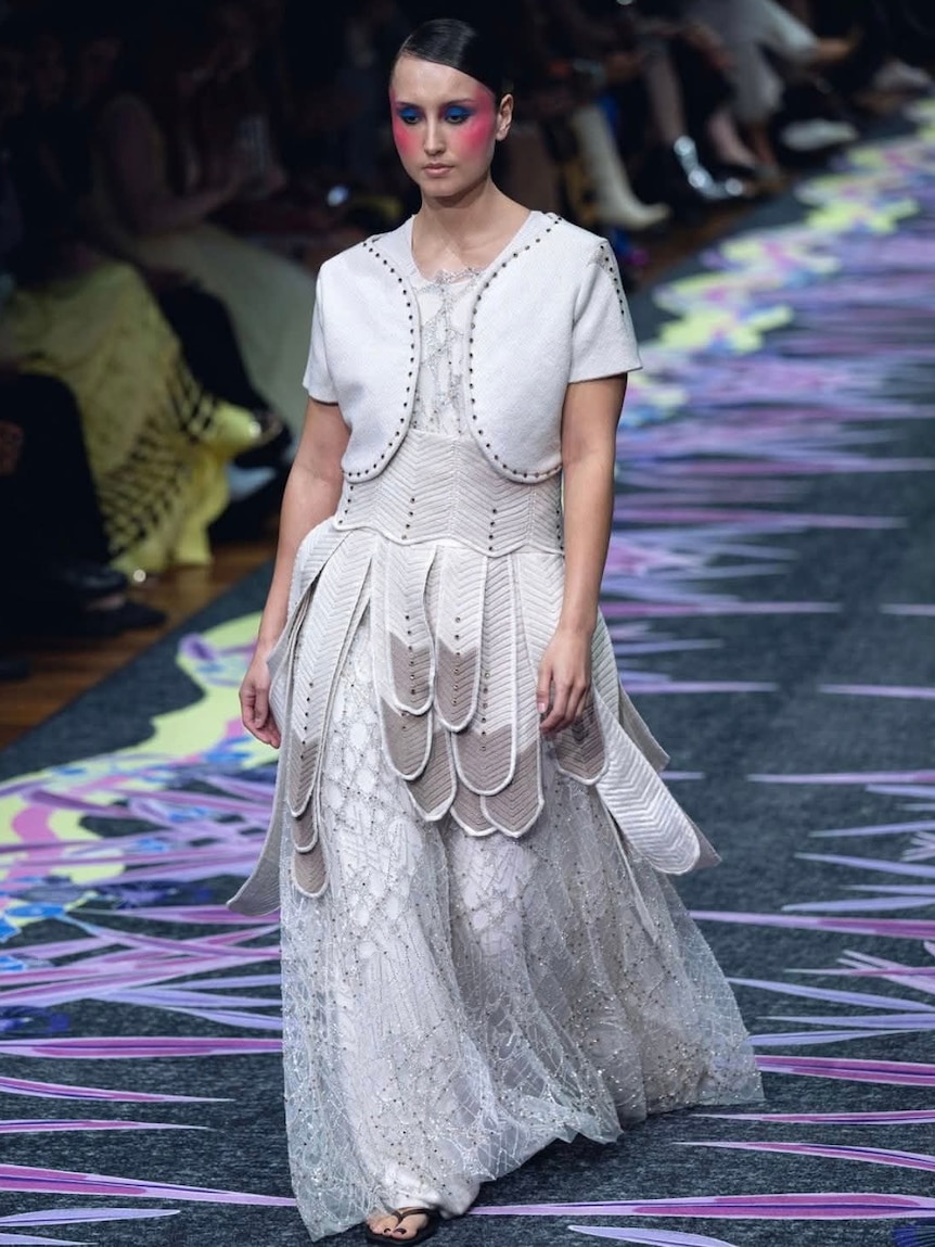 a woman walking down a runway wearing a long white dress featuring a feather waist. 