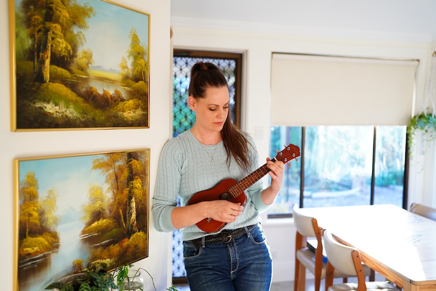 A woman standing and playing a ukelele, in a light-filled room with paintings on the wall next to her.