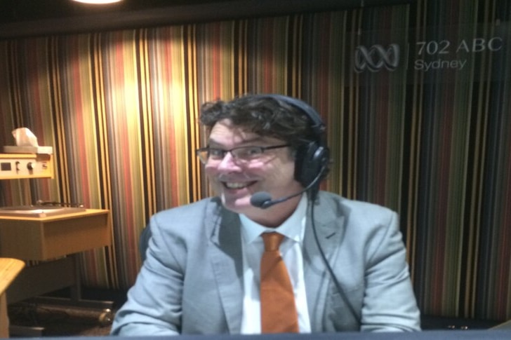 James Valentine in a blue suit and orange tie wearing a headset and sitting in a radio studio 