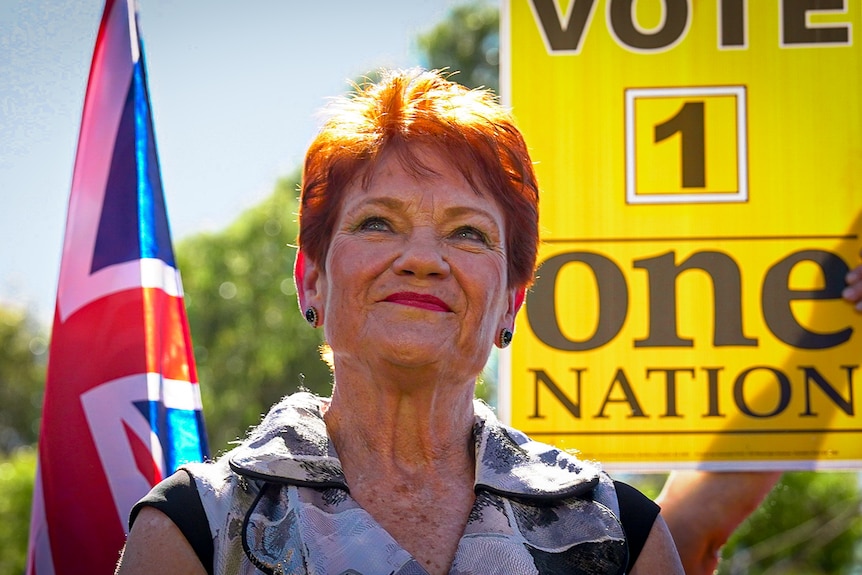 A woman with orange hair stands in front of an Australian flag and a sign saying VOTE 1 one NATION