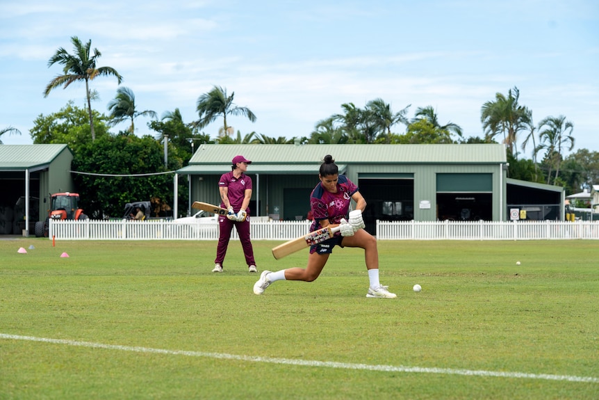 Women standing on a cricket pitch, practising batting a cricket ball