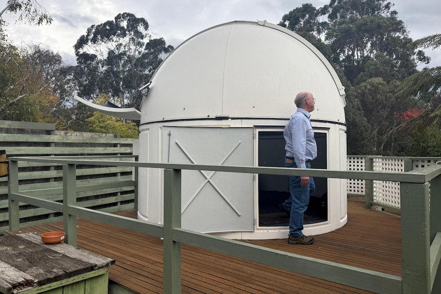 A man standing on a wooden decking, in front of a white, cylinder-shaped observatory in a backyard