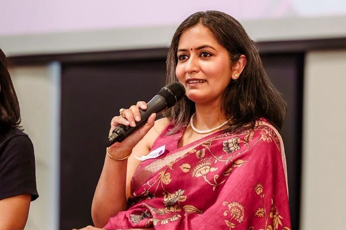A woman in a pink sari speaks into a microphone on stage as part of a panel.